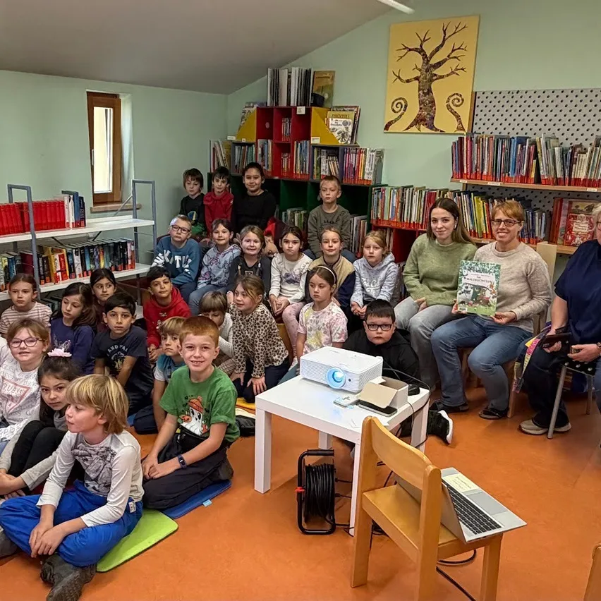 Eine Gruppe von Kindern und Erwachsenen sitzt auf dem Boden in einer Bibliothek. Eine Projektionsleinwand ist an der Wand, und eine Frau liest ein Buch.