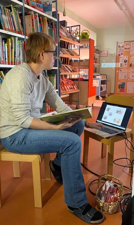Eine Person sitzt auf einem Stuhl in einer Bibliothek, liest ein Buch und benutzt einen Laptop.