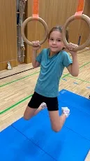 Ein junges Mädchen macht Gymnastik auf einem blauen Teppich. Sie hält zwei Holzringe mit Händen und Füßen.