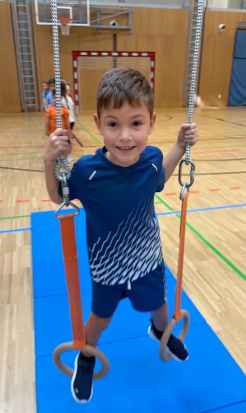 Ein junger Junge hängt an zwei Ringen auf einem blauen Teppich in einer Turnhalle.