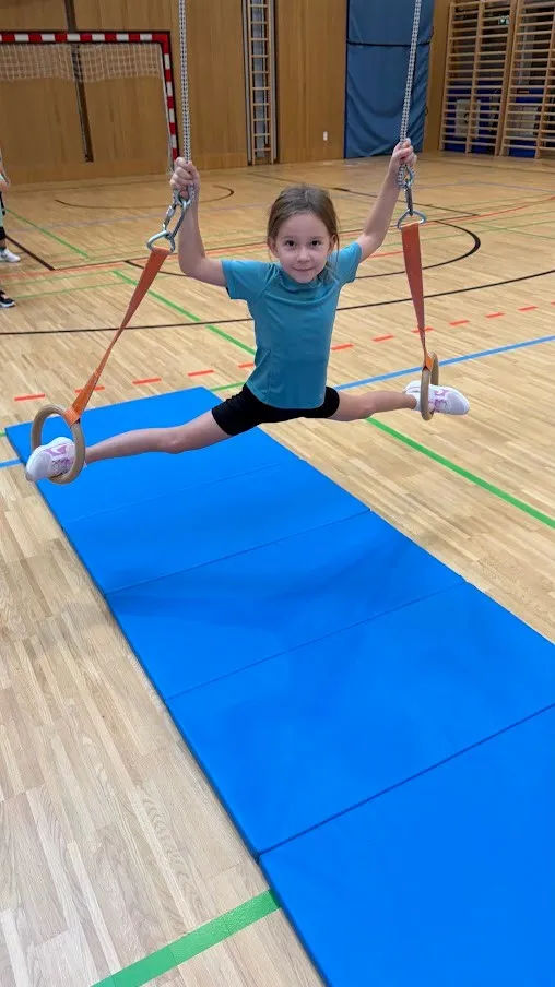 Ein kleines Mädchen schwingt auf einer Trapezstange in einer Turnhalle. Sie trägt ein blaues T-Shirt, schwarze Shorts und weiße Turnschuhe.