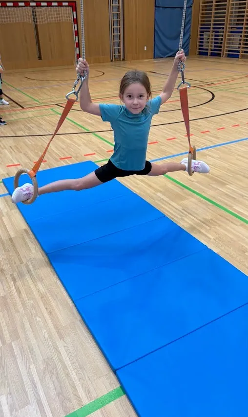 Ein kleines Mädchen schwingt auf einer Trapezstange in einer Turnhalle. Sie trägt ein blaues T-Shirt, schwarze Shorts und weiße Turnschuhe.