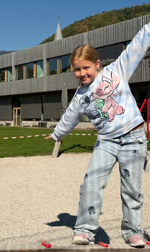 Ein kleines Mädchen steht auf einem Holzbrett im Spielplatz, trägt ein rosa Elefanten-T-Shirt und blaue Jeans mit Löchern.
