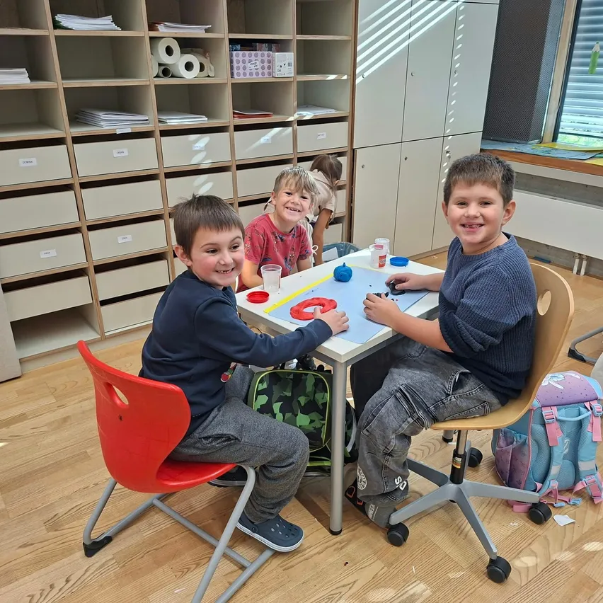 Drei junge Jungen sitzen an einem Tisch in einem Klassenzimmer. Sie lächeln und scheinen sich auf eine Aktivität zu konzentrieren. Auf dem Tisch befinden sich verschiedene Gegenstände, darunter eine blaue Matte, ein roter Becher und eine Flasche. Hinter ihnen steht ein Regal mit Büchern und anderen Gegenständen.