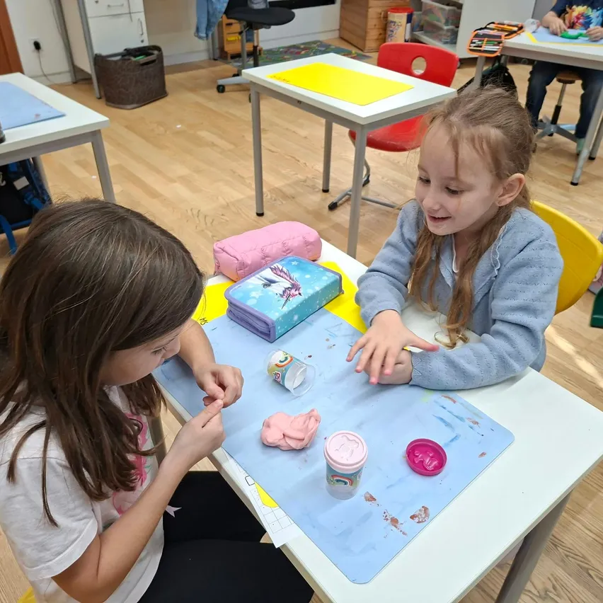 Zwei junge Mädchen sitzen an einem Tisch in einem Klassenzimmer. Ein Mädchen lächelt und schaut das andere Mädchen an, das sich auf ein Bastelprojekt konzentriert.