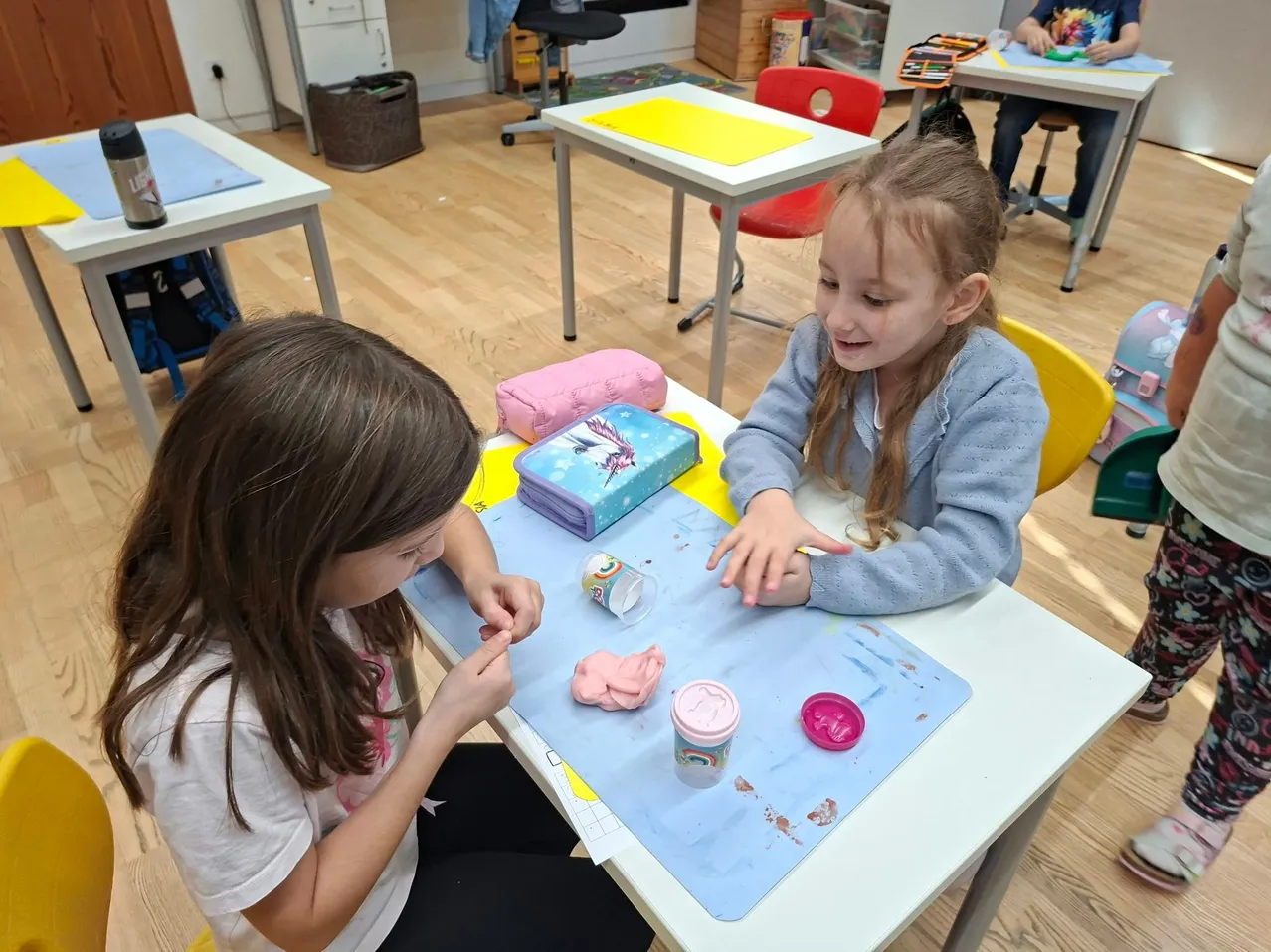 Zwei junge Mädchen sitzen an einem Tisch in einem Klassenzimmer. Ein Mädchen lächelt und schaut das andere Mädchen an, das sich auf ein Bastelprojekt konzentriert.