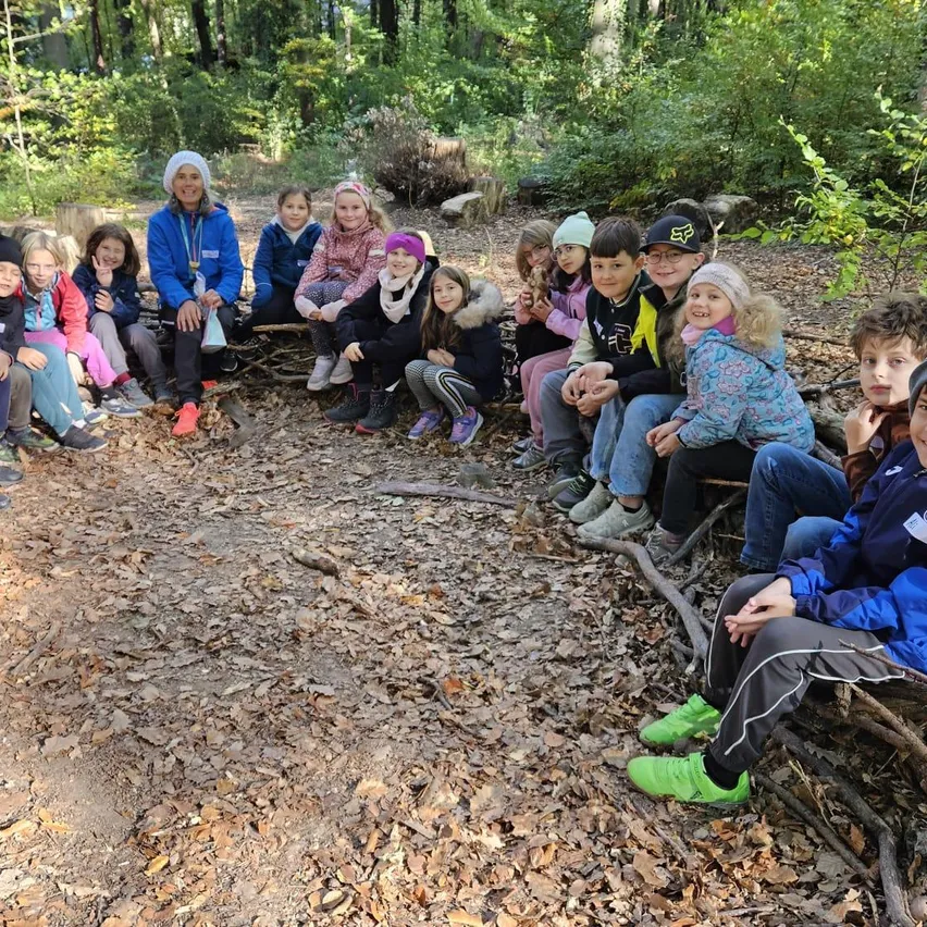 Eine Gruppe von Kindern und einer erwachsenen Frau sitzen im Kreis in einem Wald. Sie tragen alle Winterkleidung und einige von ihnen tragen Mützen.