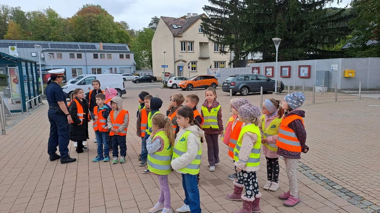 Eine Gruppe von Kindern in Sicherheitswesten steht im Kreis mit einem Polizeibeamten auf einem gepflasterten Weg.