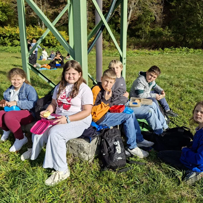 Eine Gruppe von Kindern sitzt auf einem Grasfeld, einige essen, während andere etwas betrachten.