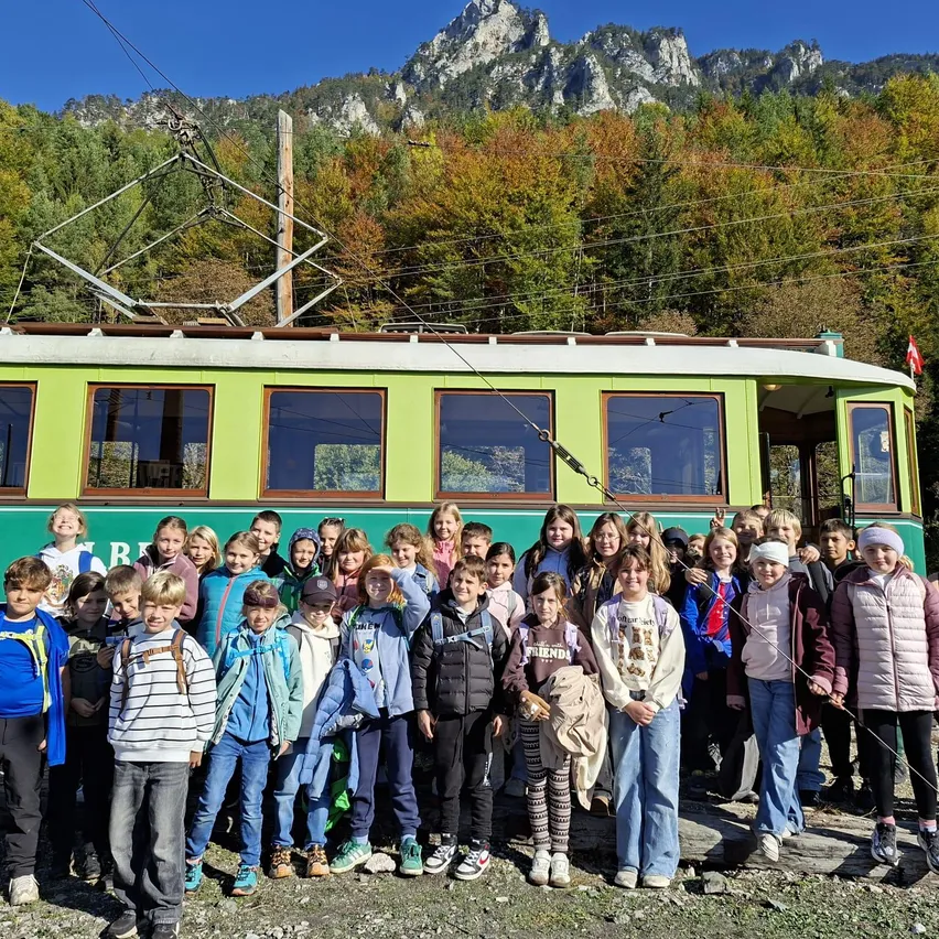 Eine Gruppe von Kindern und Erwachsenen posiert für ein Foto vor einer grünen und weißen Straßenbahn mit Bergen im Hintergrund.
