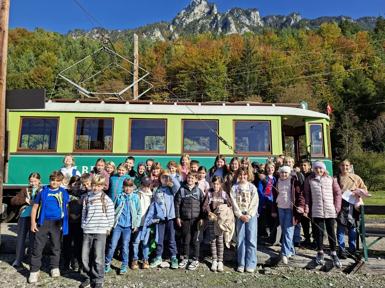 Eine Gruppe von Kindern und Erwachsenen posiert für ein Foto vor einer grünen und weißen Straßenbahn mit Bergen im Hintergrund.