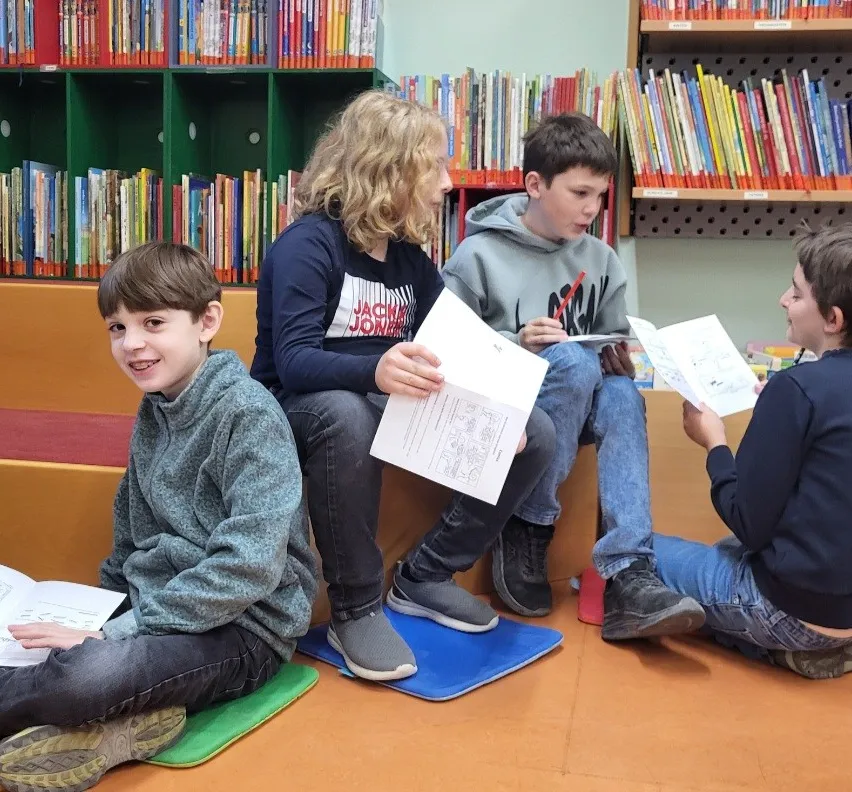 Fünf Kinder sitzen auf dem Boden in einer Bibliothek, jedes hält ein Buch und liest.