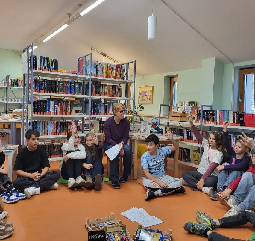 Eine Gruppe von Kindern sitzt im Kreis auf dem Boden einer Bibliothek, eine Frau steht in der Mitte und hält Papiere.