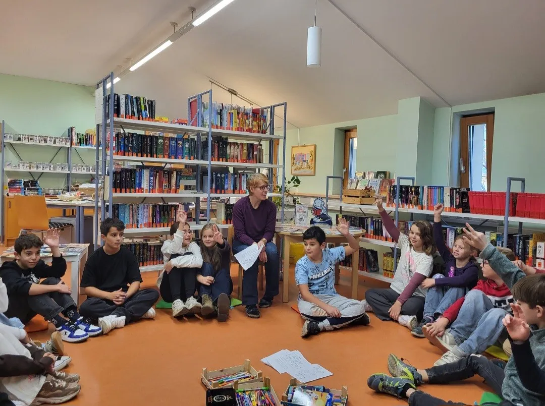Eine Gruppe von Kindern sitzt im Kreis auf dem Boden einer Bibliothek, eine Frau steht in der Mitte und hält Papiere.