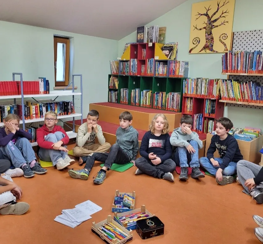 Eine Gruppe von Kindern sitzt auf dem Boden in einem Klassenzimmer, einige von ihnen tragen eine Brille und Turnschuhe. Es gibt Regale voller Bücher und ein Baumbild an der Wand.