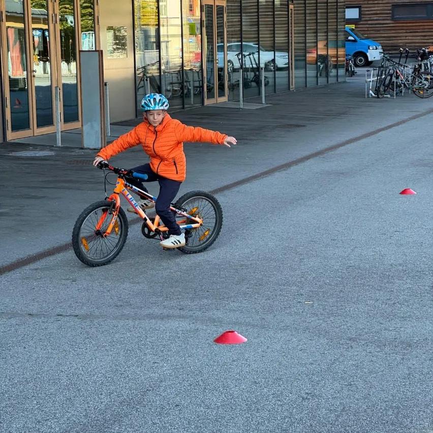 Ein junger Junge fährt auf einer asphaltierten Straße mit einem Fahrrad. Er trägt einen Helm und eine orangefarbene Jacke.