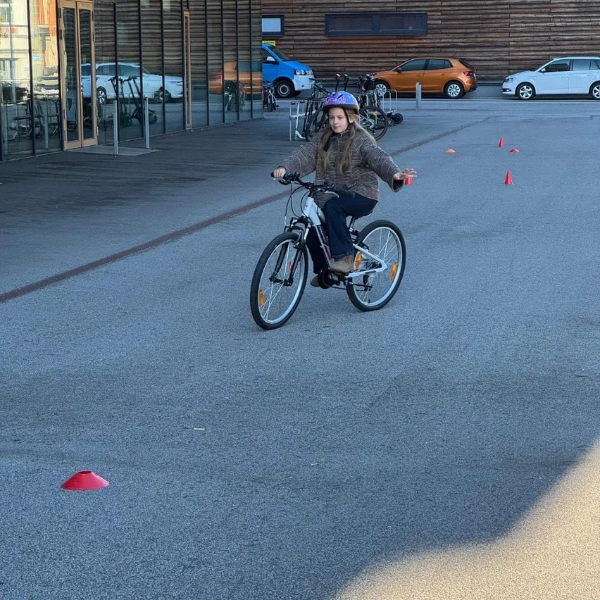Ein junges Mädchen fährt auf einem gepflasterten Weg mit einem Fahrrad. Um sie herum sind Verkehrskegel platziert. Hinter ihr befindet sich ein Gebäude mit Glaswänden und einer hölzernen Wand.