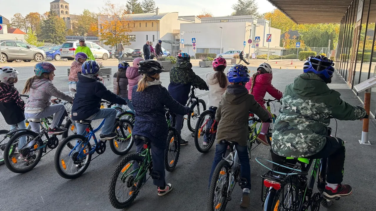 Eine Gruppe von Kindern mit Helmen fährt auf Fahrrädern in einem Parkplatz, mit geparkten Autos und einem Gebäude im Hintergrund.