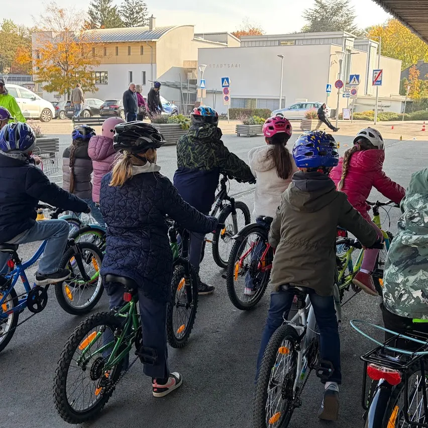Eine Gruppe von Kindern mit Helmen fährt auf Fahrrädern in einem Parkplatz, mit geparkten Autos und einem Gebäude im Hintergrund.