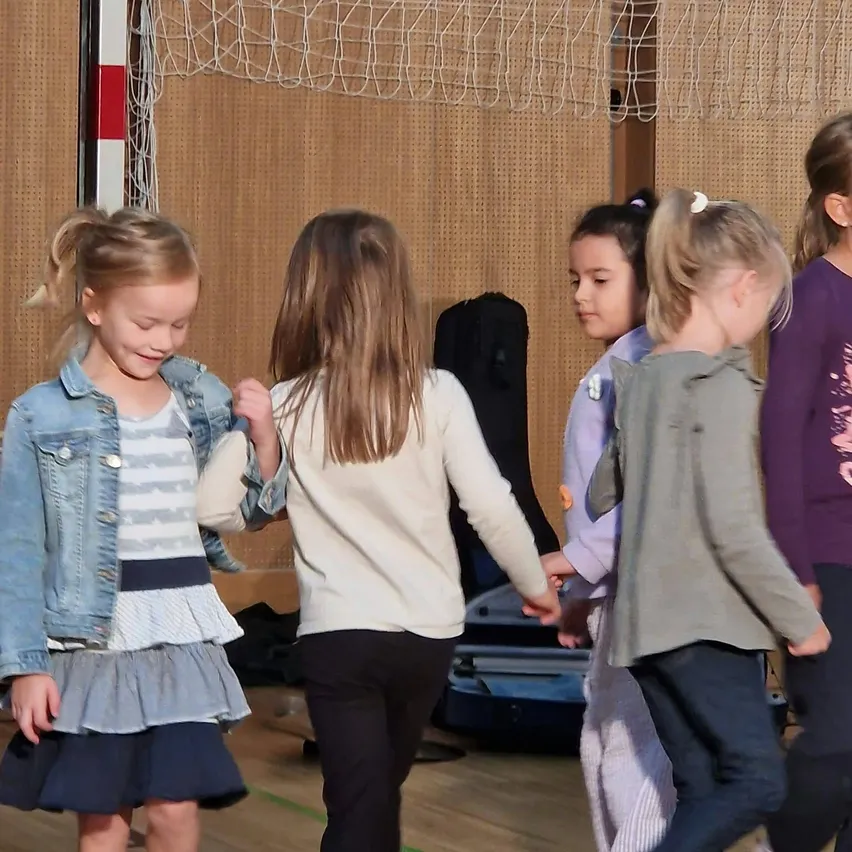Fünf junge Mädchen spielen ein Spiel in einer Turnhalle. Das Mädchen auf der linken Seite trägt eine Jeansjacke und einen Rock, während die anderen langärmelige Hemden und Hosen tragen.