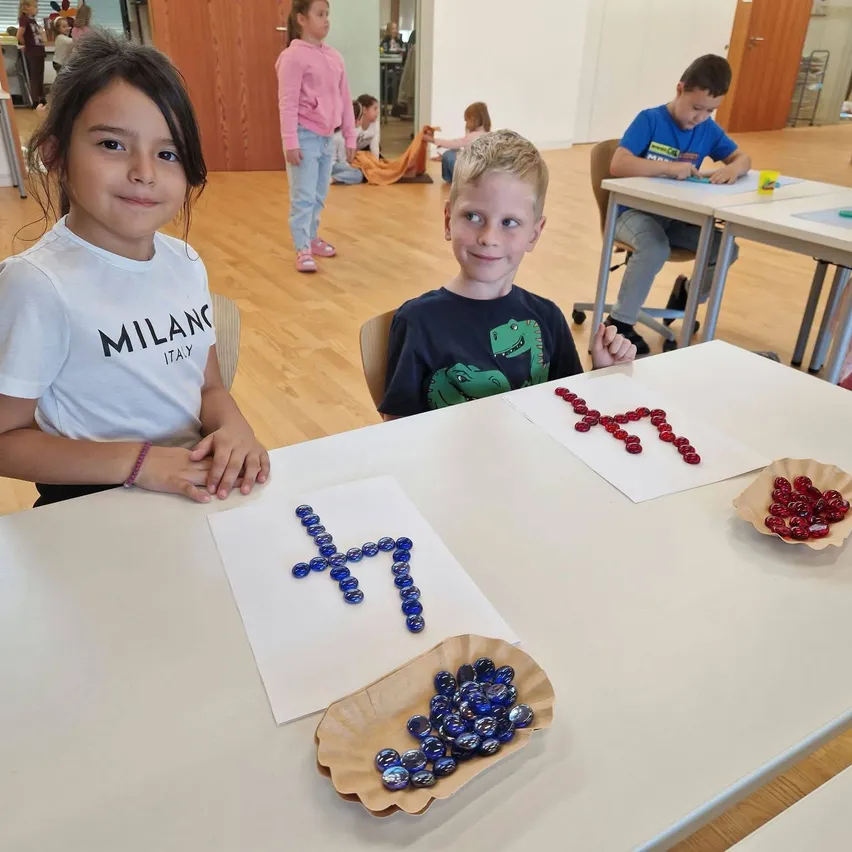 Zwei Kinder, ein Mädchen und ein Junge, sitzen an einem Tisch im Klassenzimmer. Das Mädchen trägt ein weißes T-Shirt mit dem Wort 'Milan' darauf und der Junge trägt ein blaues T-Shirt mit einem Dinosaurier darauf. Sie lächeln beide und schauen in die Kamera.