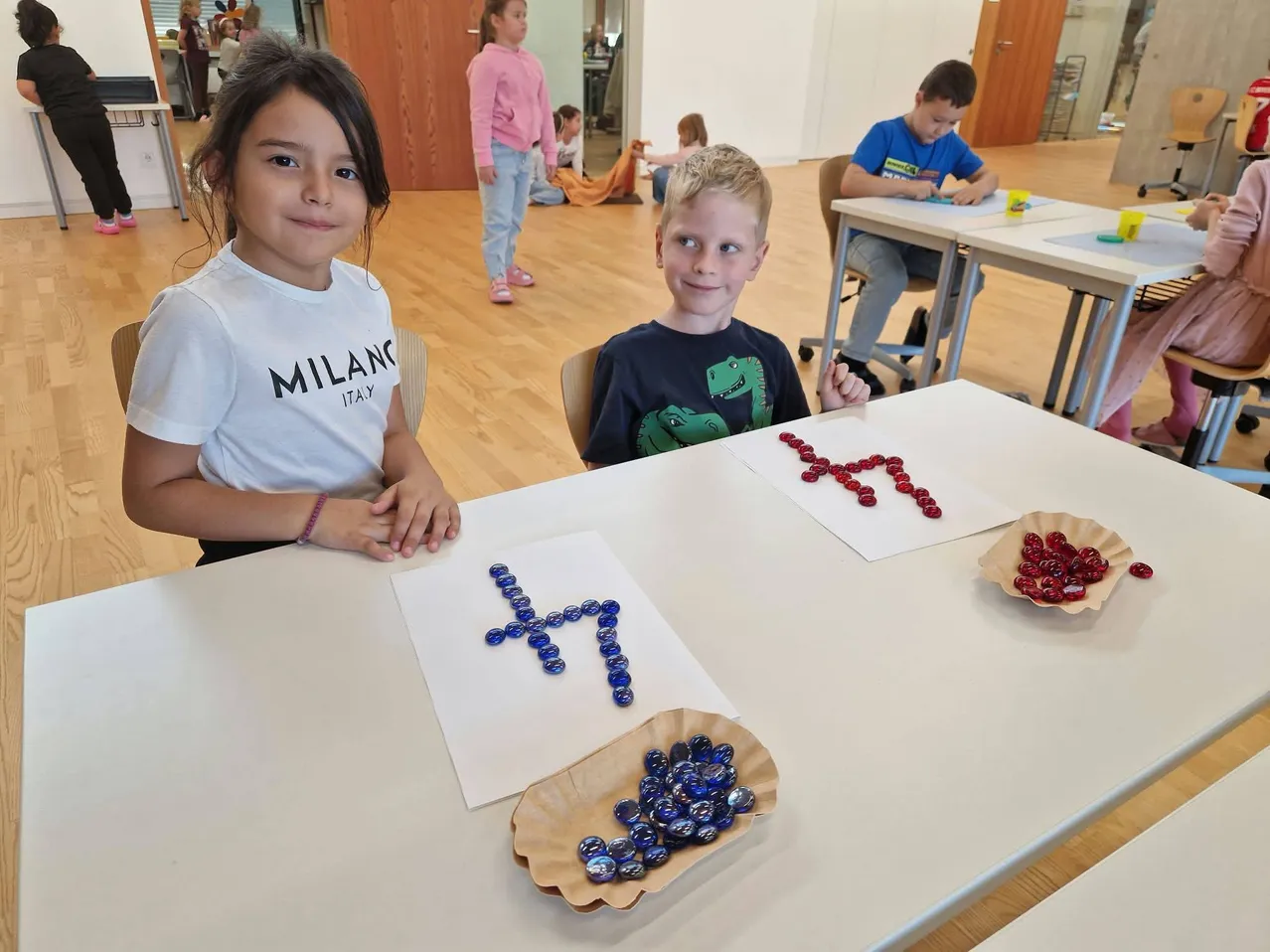 Zwei Kinder, ein Mädchen und ein Junge, sitzen an einem Tisch im Klassenzimmer. Das Mädchen trägt ein weißes T-Shirt mit dem Wort 'Milan' darauf und der Junge trägt ein blaues T-Shirt mit einem Dinosaurier darauf. Sie lächeln beide und schauen in die Kamera.