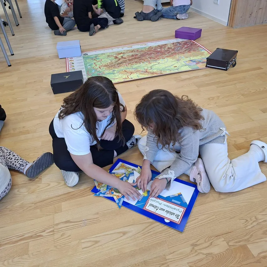 Zwei junge Mädchen sitzen auf dem Boden und beschäftigen sich mit einer Puzzle-Aktivität. Sie sind von anderen Kindern umgeben, die entweder auf dem Boden oder auf Stühlen sitzen. Ein großer Plan liegt auf dem Boden und verschiedene Gegenstände sind verstreut.