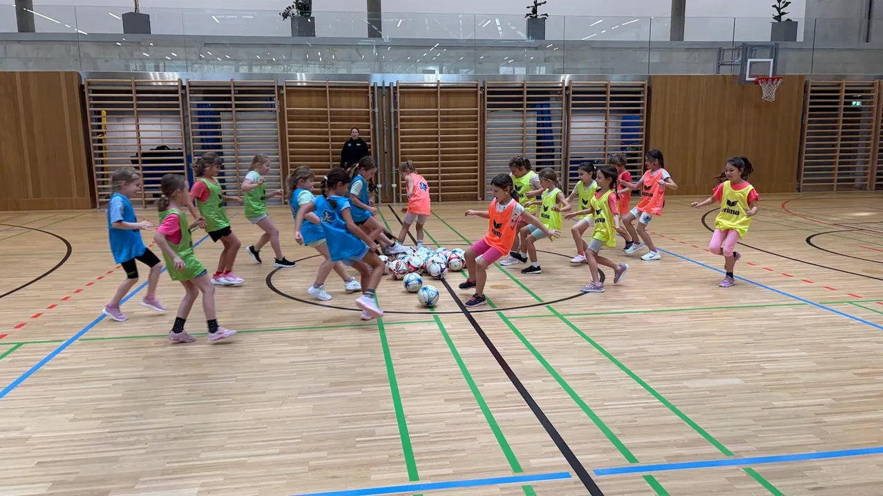 Eine Gruppe junger Mädchen in passenden Uniformen spielt Fußball auf einem Hallenplatz. Sie treten und passen den Ball aneinander.