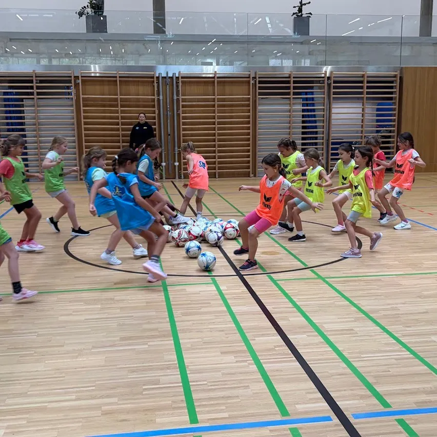 Eine Gruppe junger Mädchen in passenden Uniformen spielt Fußball auf einem Hallenplatz. Sie treten und passen den Ball aneinander.