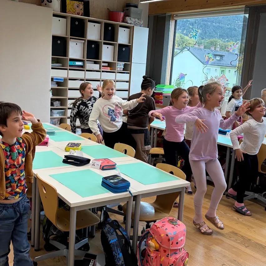 Eine Gruppe von Kindern in einem Klassenzimmer tanzt und winkt mit den Händen, während sie Freizeitkleidung tragen.