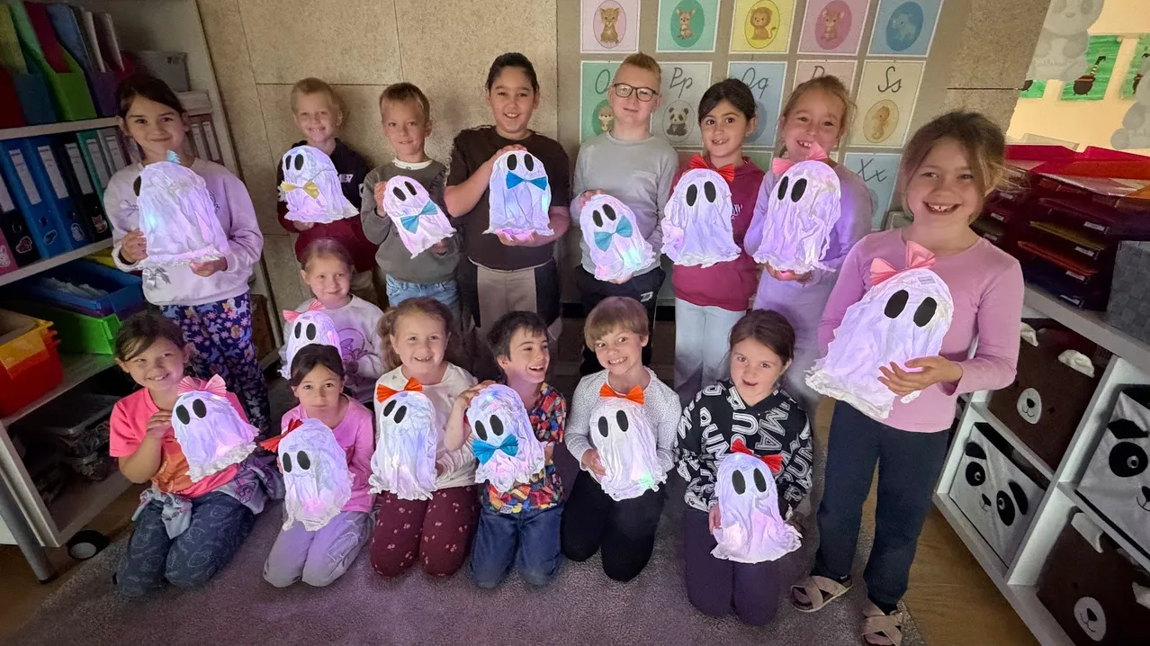 Eine Gruppe von Kindern in einem Klassenzimmer hält selbstgemachte Geistermasken hoch und lächelt für ein Foto.