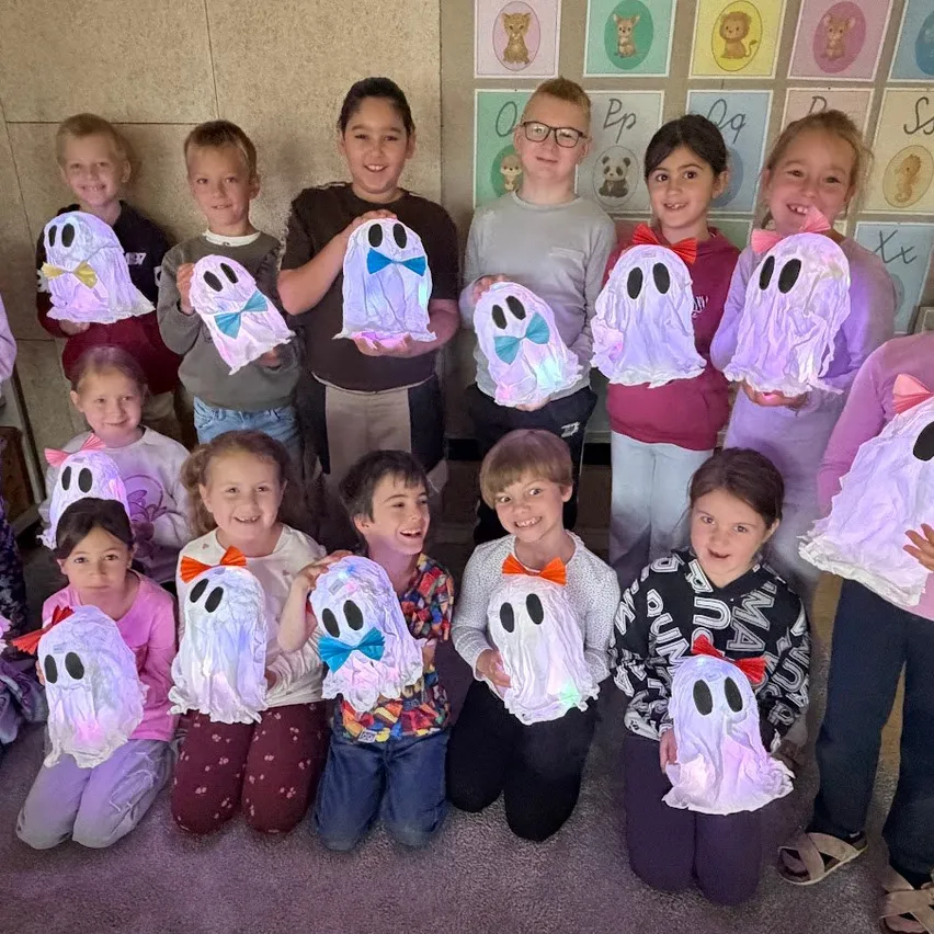 Eine Gruppe von Kindern in einem Klassenzimmer hält selbstgemachte Geistermasken hoch und lächelt für ein Foto.