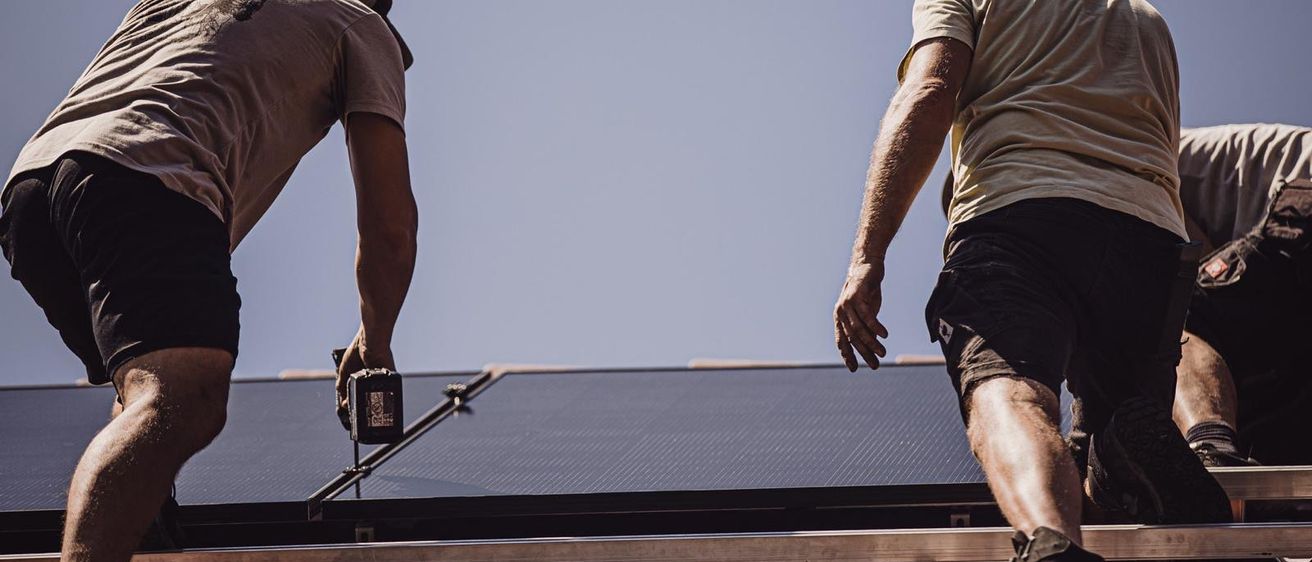 Zwei Männer installieren Solarmodule auf einem Dach. Ein Mann hält ein Werkzeug, während der andere in der Nähe steht. Der Himmel ist klar und blau.