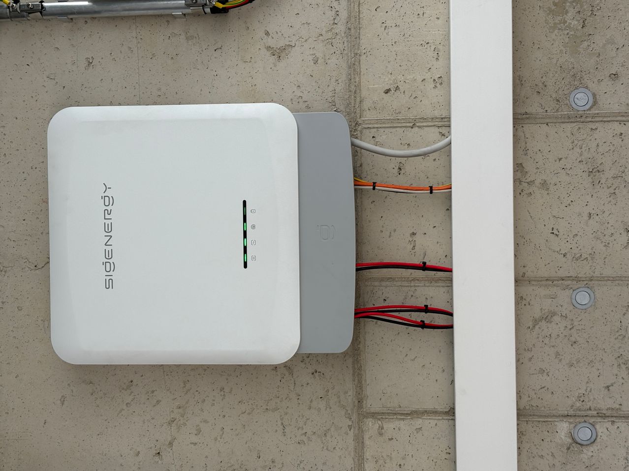 Ein weißes und graues Gerät mit der Aufschrift Signenergy ist an einer Betonwand mit roten und weißen Kabeln montiert.
