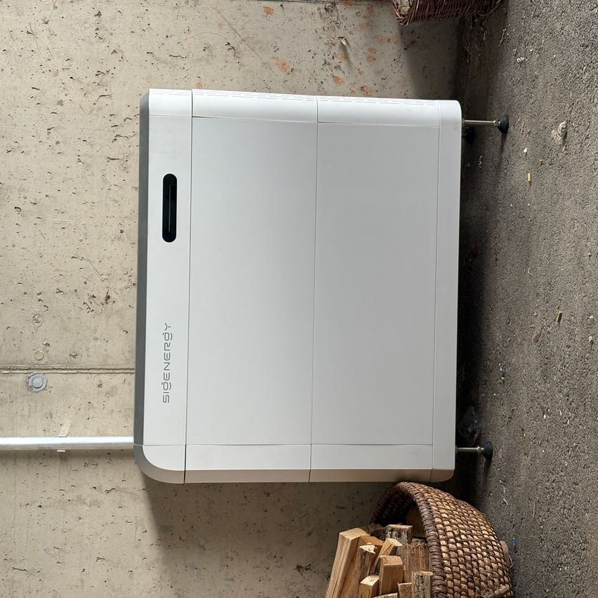 Ein weißer, rechteckiger Sidenergy-Heizer ist an einer Betonwand montiert. Er hat einen kleinen schwarzen Griff an der Seite. Neben ihm steht ein Korb mit Holzscheiten.