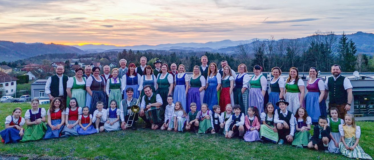 Eine Gruppe von Menschen in traditioneller Kleidung, einschließlich Kinder, posiert für ein Foto auf einem Rasenfeld mit Bergen im Hintergrund.