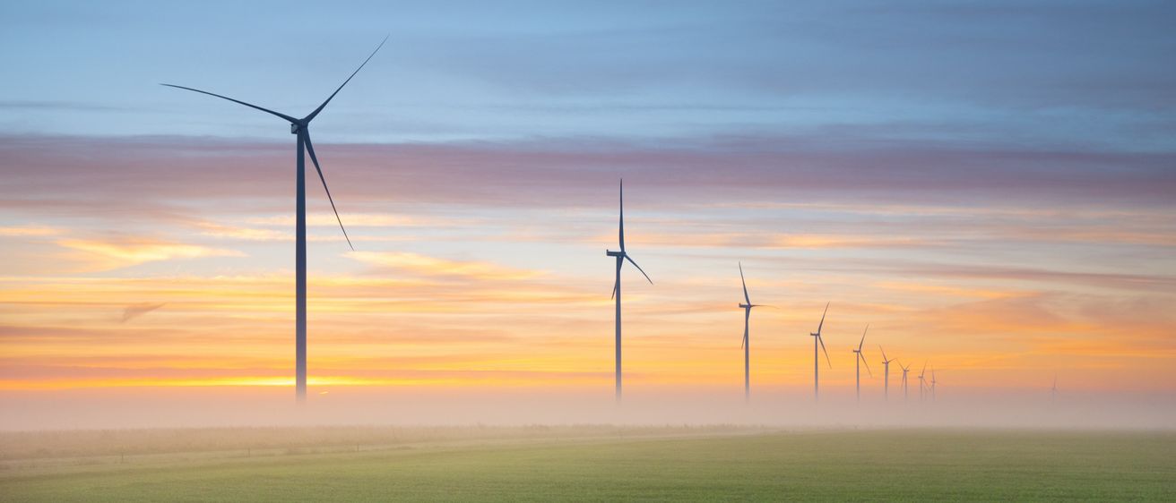 Ein Feld mit Windrädern steht in einer nebligen Landschaft, mit der untergehenden Sonne hinter einem Horizont von Wolken.