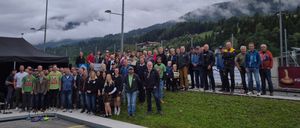 Eine Gruppe von Menschen in Winterkleidung steht auf einer Betonplattform vor einem Rasenbereich mit Zaun und Bergen im Hintergrund.