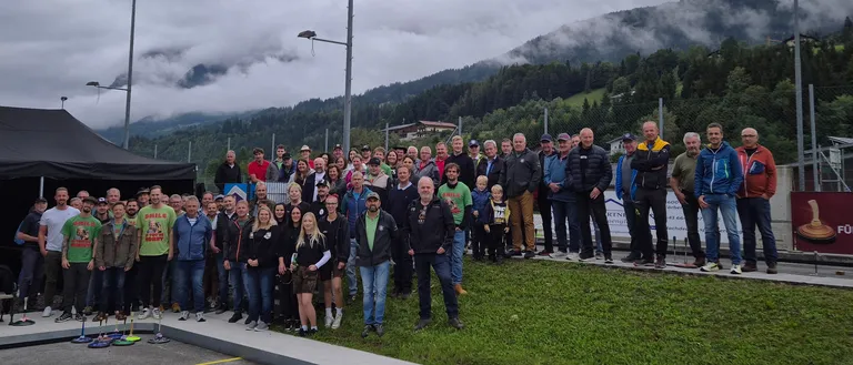 Eine Gruppe von Menschen in Winterkleidung steht auf einer Betonplattform vor einem Rasenbereich mit Zaun und Bergen im Hintergrund.