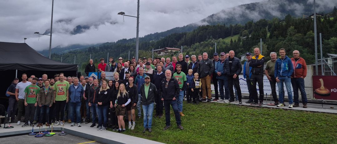 Eine Gruppe von Menschen in Winterkleidung steht auf einer Betonplattform vor einem Rasenbereich mit Zaun und Bergen im Hintergrund.