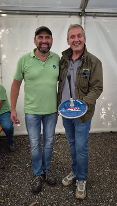 Zwei Männer stehen eng beieinander und lächeln, einer hält eine runde blaue Plakette mit 'Salzburg' darauf. Einer trägt ein grünes Poloshirt mit Logo, der andere eine grüne Jacke mit einem Handy in der Tasche.