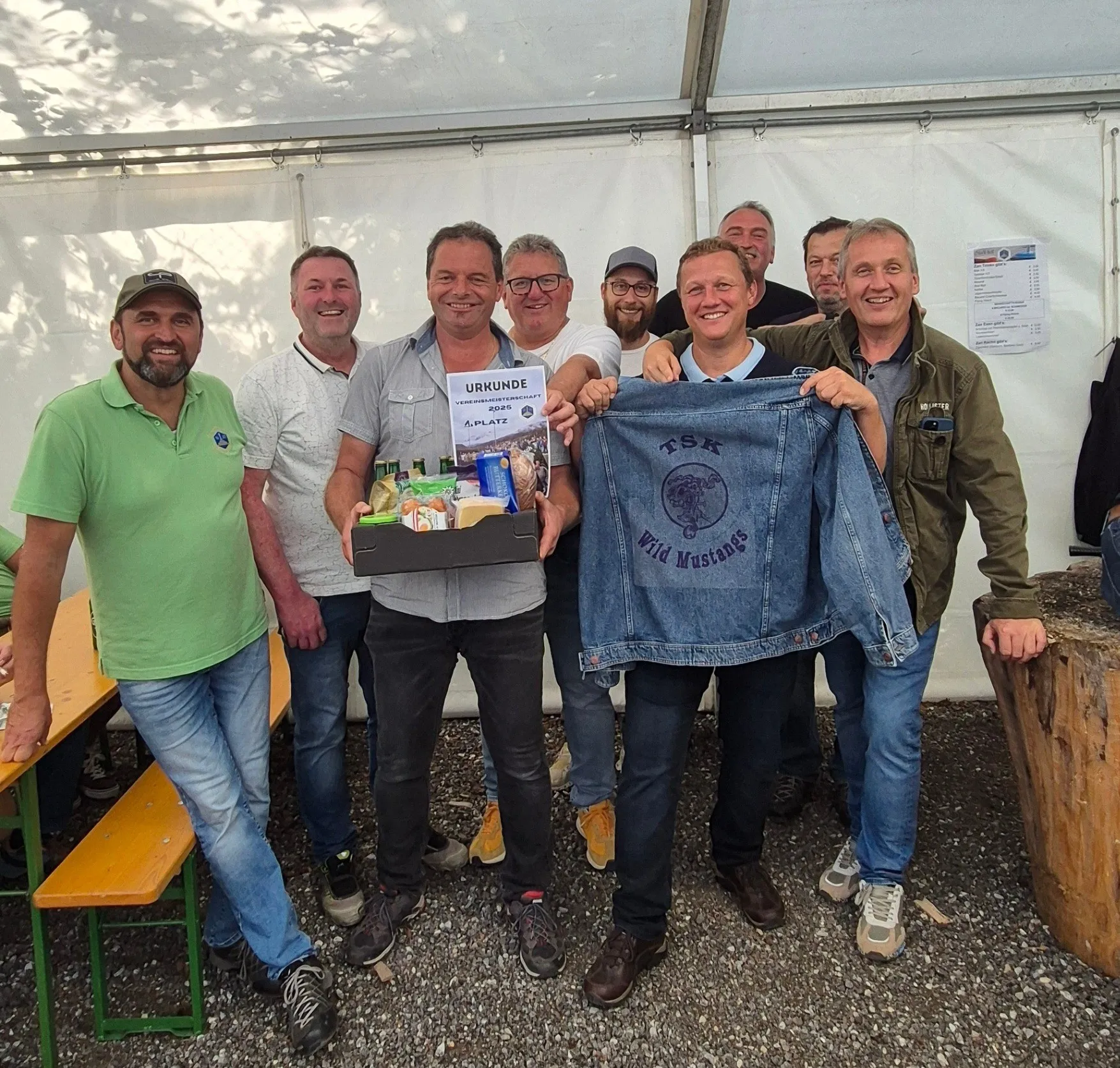 Eine Gruppe von Männern steht lächelnd zusammen in einem Zelt. Ein Mann hält eine Jeansjacke und eine Box. Sie sind wahrscheinlich bei einer Veranstaltung oder einem Treffen.