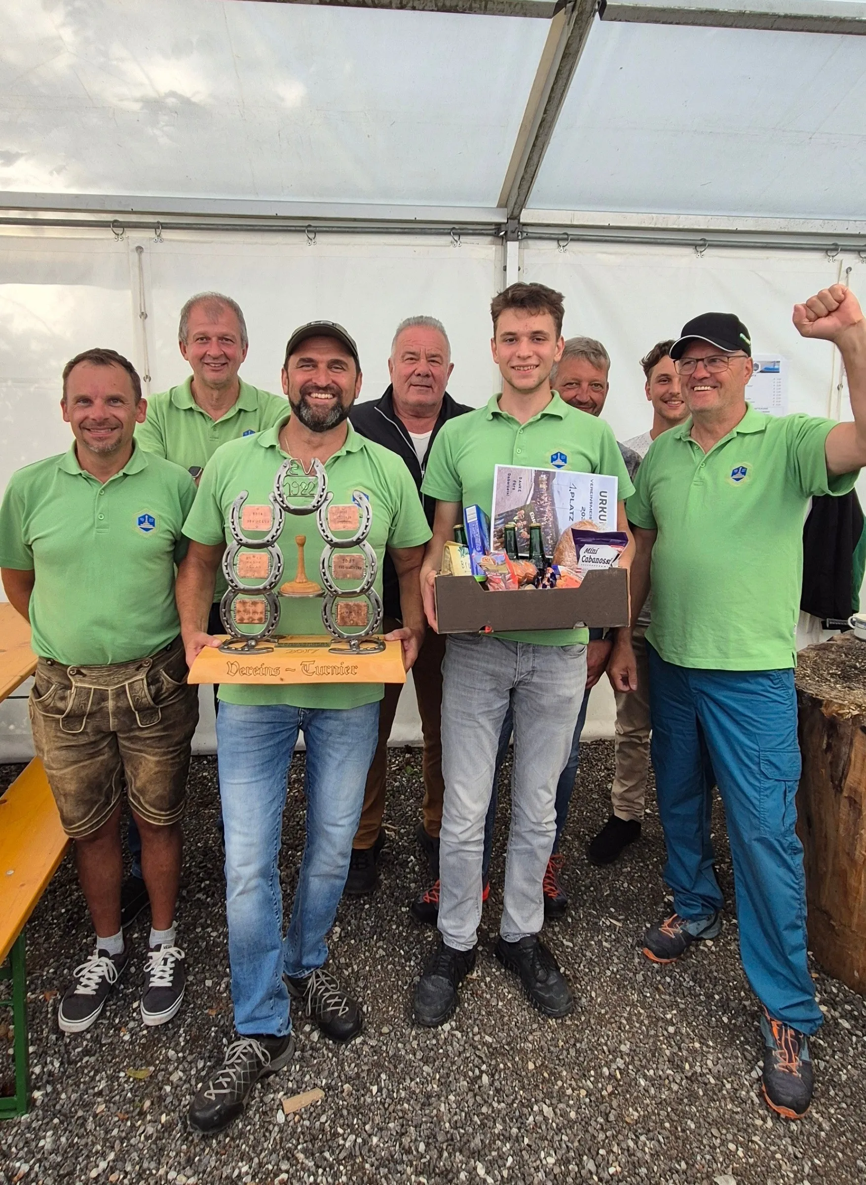 Eine Gruppe von Männern in grünen Poloshirts, einige mit Mützen, stehen in einer Reihe. Der Mann in der Mitte hält einen Pokal und eine Box. Sie befinden sich in einer Außenumgebung mit einer Holzbank.