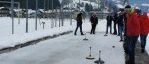 Eine Gruppe von Menschen spielt ein Curlingspiel auf einer Eisbahn mit verschneigten Bergen im Hintergrund.