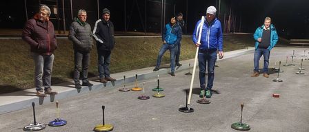 Eine Gruppe von Menschen spielt nachts draußen ein Spiel. Sie stehen um einen kreisförmigen Platz mit farbigen Markierungen. Ein Mann in einer blauen Jacke hält einen Stock.