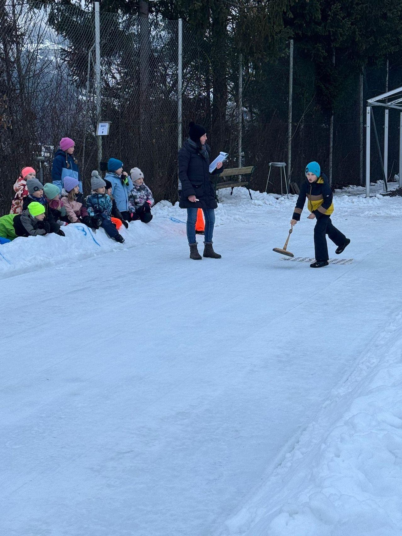 Ein Kind übt Curling auf einer verschneiten Eisbahn, während eine Frau eine Clipboard hält und eine Gruppe von Kindern zuschaut. Bäume und ein Zaun sind im Hintergrund.