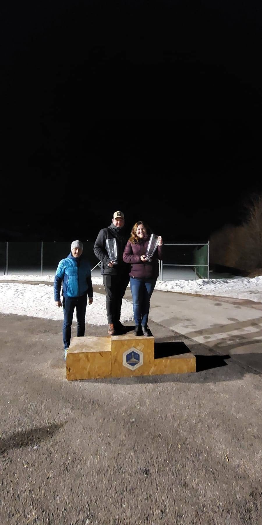 Three people stand on a podium at night, holding trophies. A man in a blue jacket stands to the left, a smiling woman in a purple jacket stands to the right, and a man in a hat stands in the middle. Snow covers the ground.