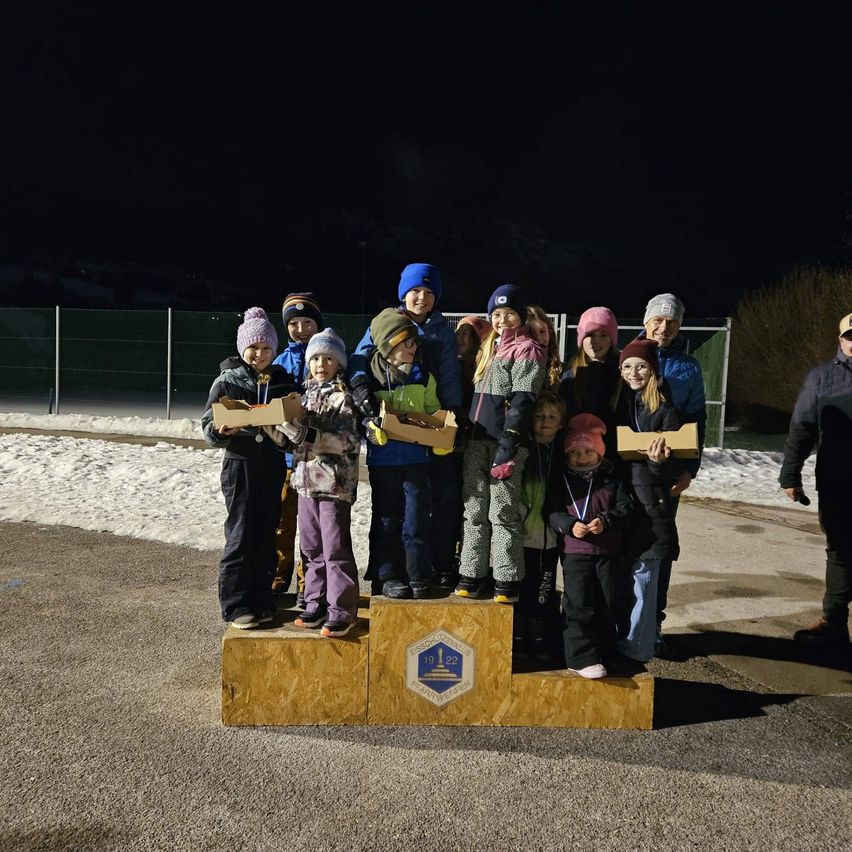 A group of children and adults stand on a podium at night, holding boxes, with a logo and a snowy ground in the background.