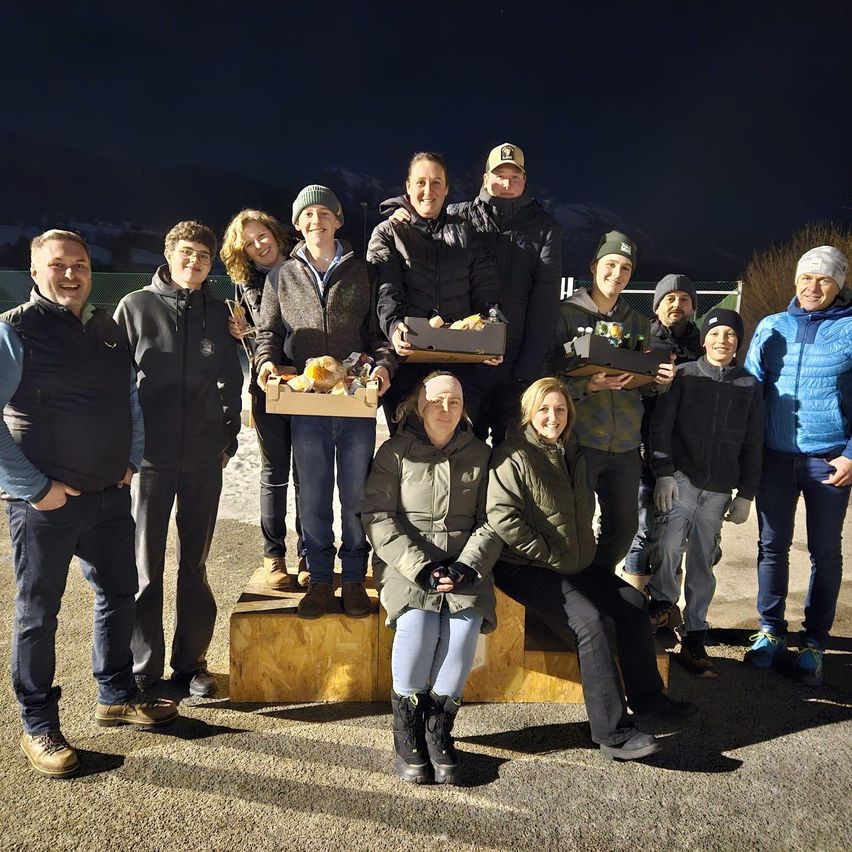 A group of people dressed warmly pose for a photo at night, some sitting, some standing, holding boxes of food.