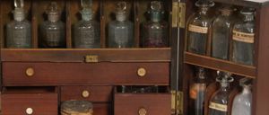 Ein Vintage-Medizinkasten aus Holz mit Messingbeschlägen, der verschiedene Glasflaschen und Gläser mit Etiketten enthält, in mehreren Schubladen und Fächern angeordnet, auf einer weißen Oberfläche platziert.